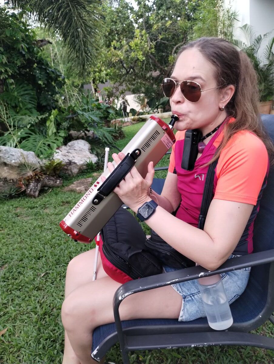 Nina outdoors in sunglasses, playing a Hammond-style melodica, surrounded by greenery